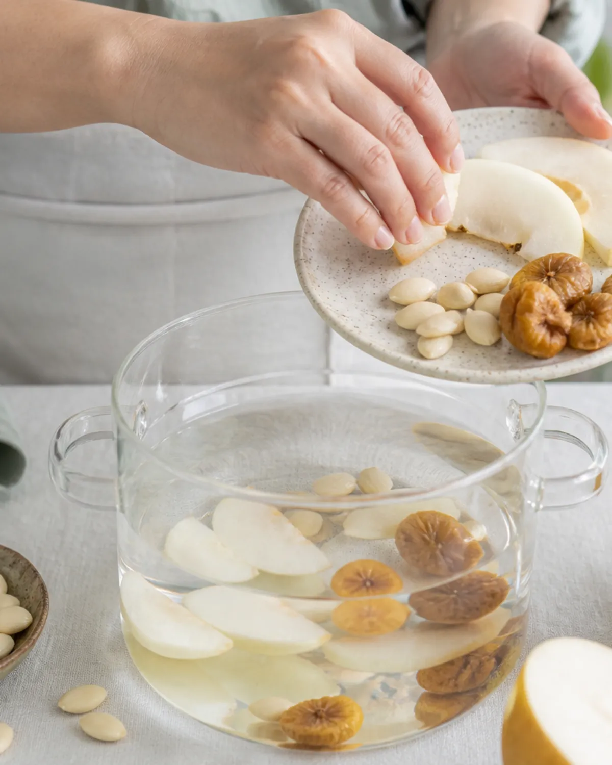 Hands adding snow pear slices and apricot kernels to a soup pot
