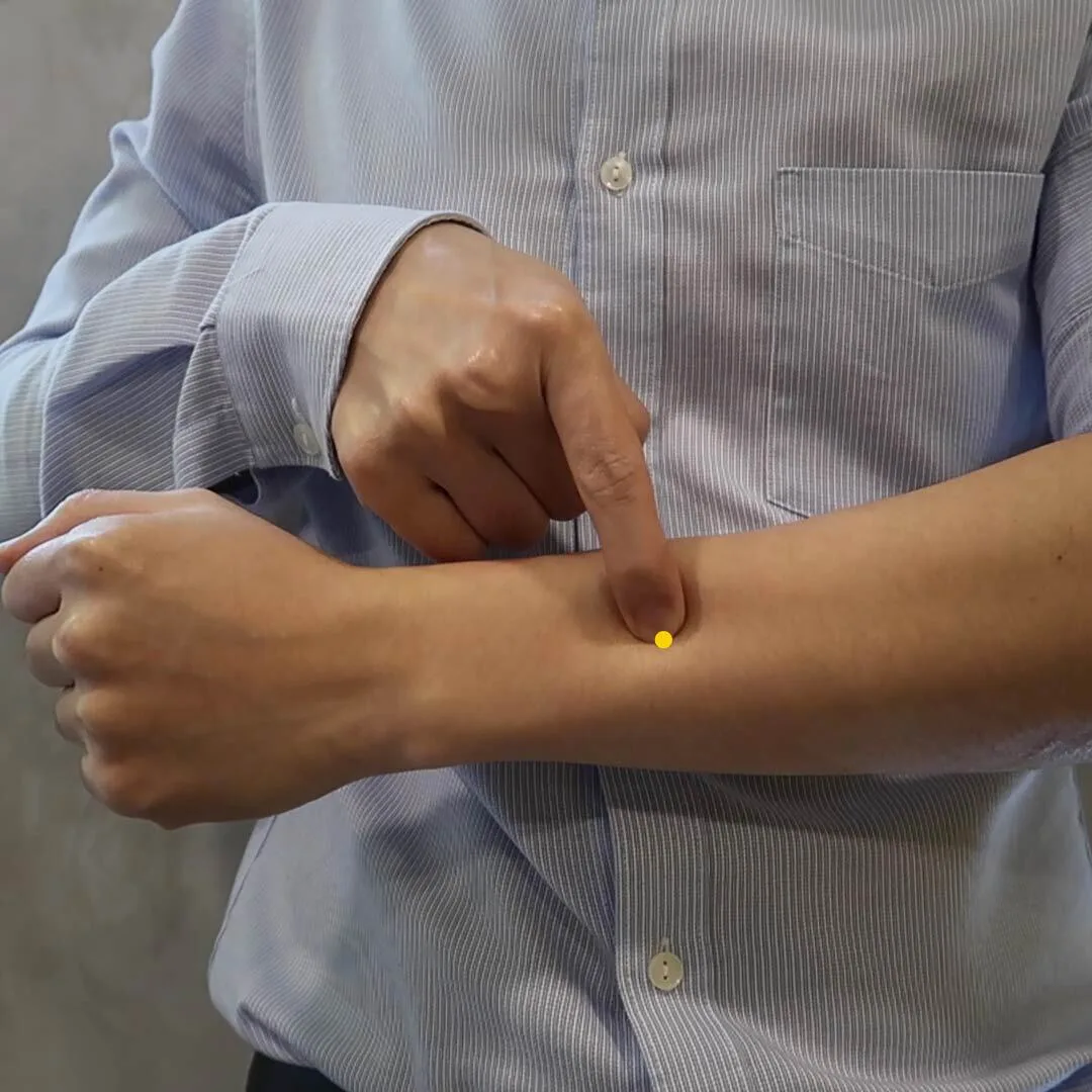 Index finger pointing to Zhi Gou SJ6 on the outer forearm, marked with a yellow dot about three cun above the wrist crease