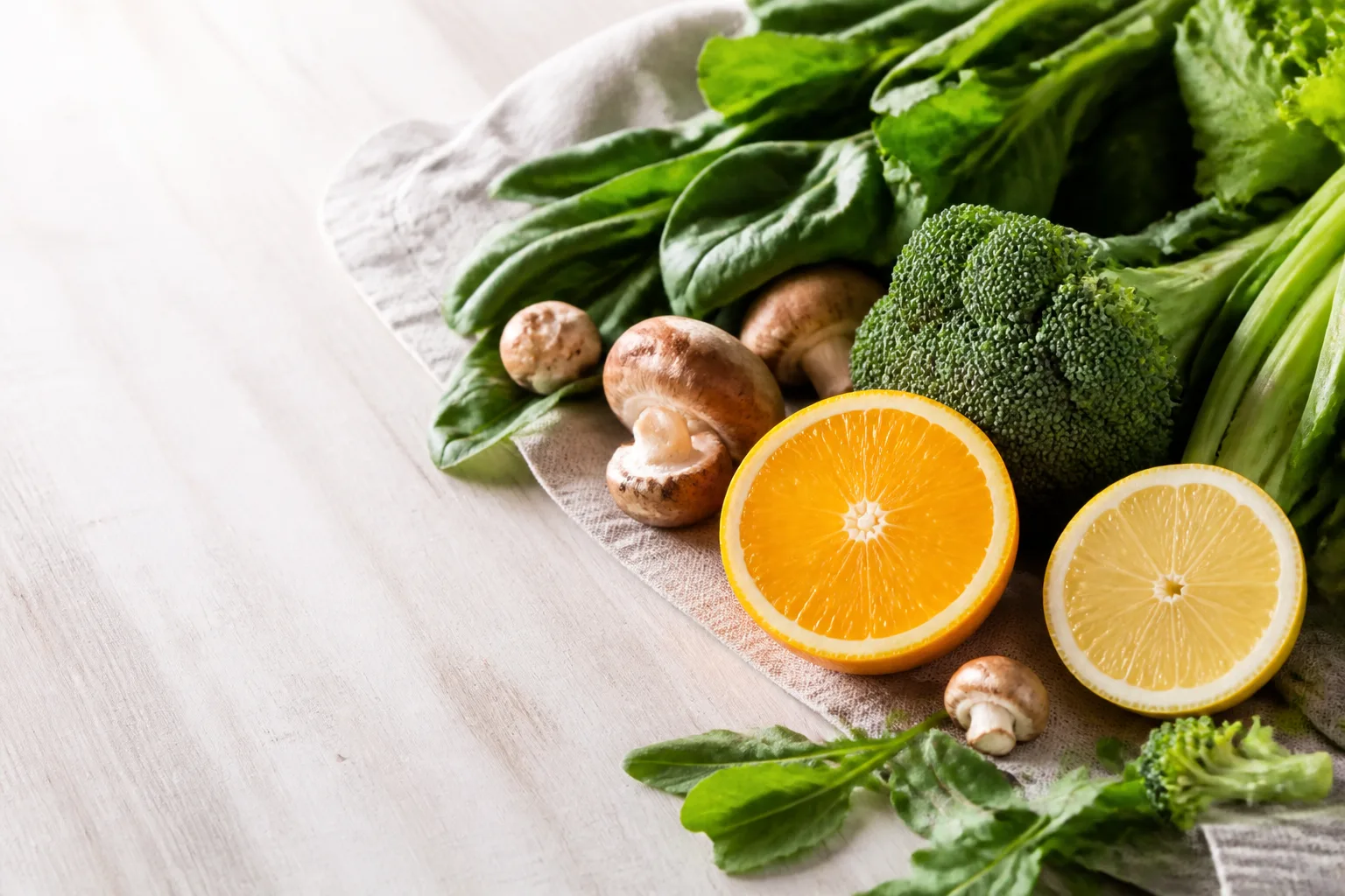 Fresh leafy greens, citrus, and mushrooms on a pale board in morning light