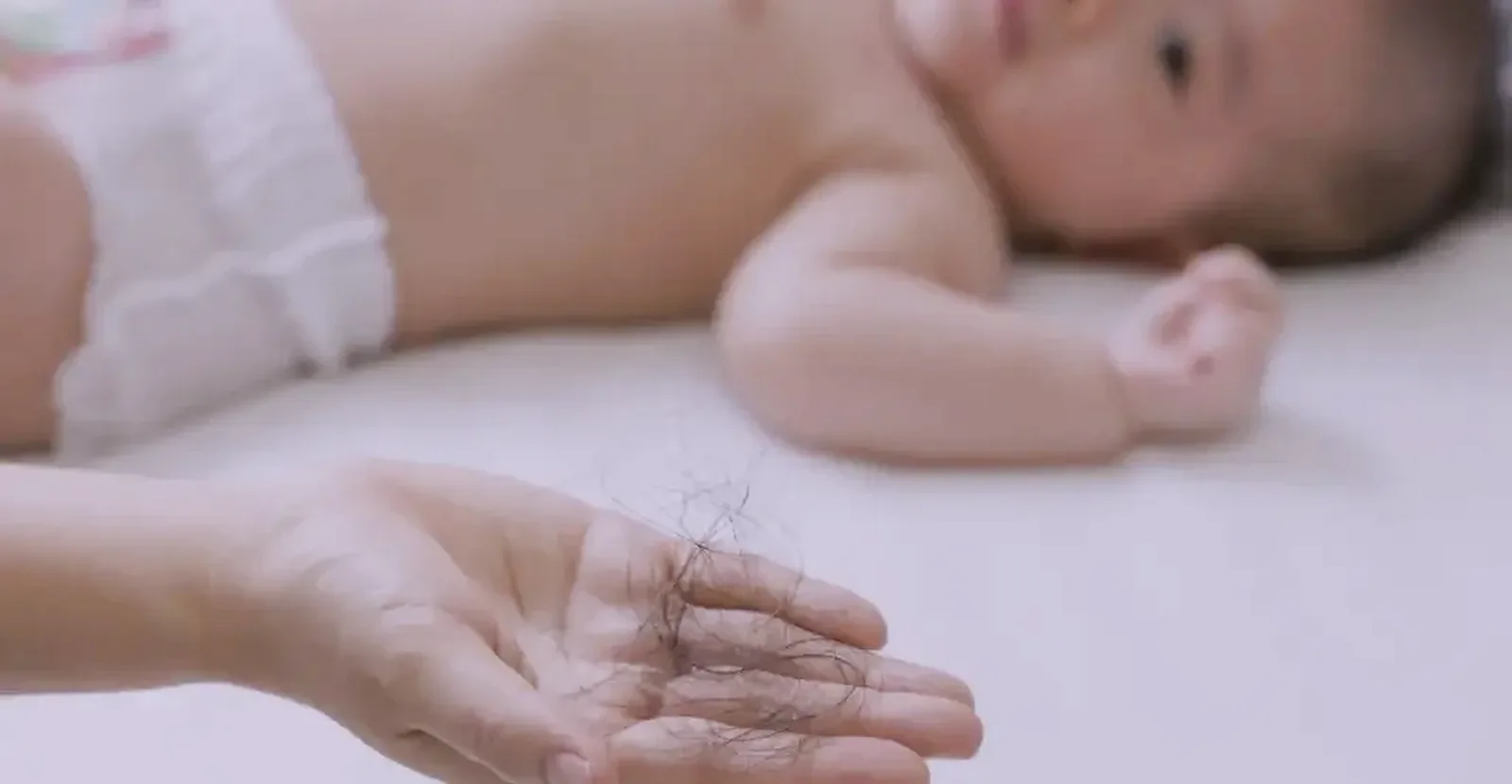 Hand holding shed postpartum hair with newborn soft-focus in the background
